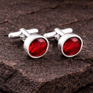 Può includere: Un paio di gemelli argentati con grandi pietre preziose rosse, sfaccettate e di forma ovale. I gemelli sono posti su uno sfondo scuro e strutturato, evidenziando il colore vibrante e la lucentezza delle pietre. Accessori per abbigliamento formale.