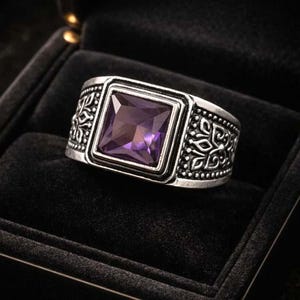 May include: A silver-toned ring with a square, purple gemstone set in the center. The ring has an ornate, engraved design around the band. The ring is displayed in a black jewelry box.