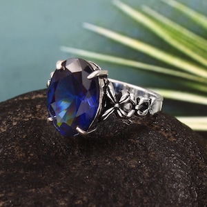 May include: A silver ring with an oval, iridescent blue gemstone. The band has floral patterns. The ring is set on a dark, textured surface with a blurred green and white background. The gemstone's dimensions are approximately 2 cm.