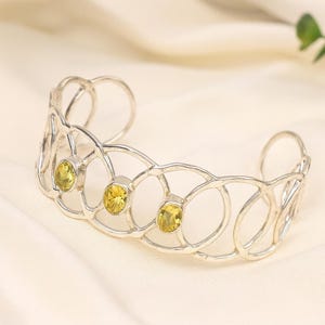 Op de afbeelding: Een zilverkleurige manchetarmband met een open ontwerp met drie ovale gele edelstenen. De armband heeft een geometrisch patroon van onderling verbonden cirkels en wordt gepresenteerd op een crèmekleurig oppervlak.