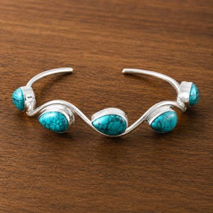 May include: A silver cuff bracelet featuring a wavy, open-ended band design. The bracelet is set with four teardrop-shaped turquoise stones, each displaying a distinct dark matrix pattern, arranged along the undulating silver metal frame.