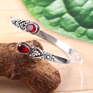 May include: A silver cuff bracelet with two teardrop-shaped, deep red gemstones. The bracelet has detailed metalwork around the gemstones and along the band, giving a vintage or bohemian look. The bracelet is displayed on a wooden surface.