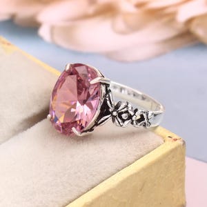 Könnte beinhalten: Ein silberner Ring mit einem großen, oval geschliffenen rosa Edelstein. Das Ringband ist mit floralen Details an der Seite verziert. Der Edelstein ist der Blickfang, der das Licht einfängt und seine Facetten zur Geltung bringt. Ein klassisches und elegantes Schmuckstück.