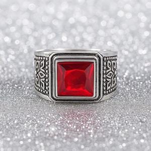 May include: A silver ring with a large, square, red gemstone. The band has an ornate, repeating pattern. The ring is set against a sparkly, silver background.