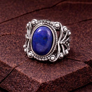 Puede incluir: Un anillo de plata con una piedra preciosa ovalada de color azul oscuro. El anillo tiene un diseño ornamentado con elementos decorativos y el texto "FizaJewels". La piedra preciosa es el punto focal, engastada en un engaste detallado de estilo vintage.