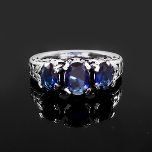 Op de afbeelding: Een zilveren ring met drie ovale, diepblauwe edelstenen. De ring heeft een sierlijk, bloemachtig ontwerp op de band. De edelstenen zijn in een rij geplaatst, wat een klassieke en elegante uitstraling creëert. De ring wordt gepresenteerd op een reflecterend zwart oppervlak.