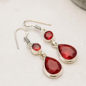 Può includere: Orecchini pendenti in argento con pietre preziose rosse. Ogni orecchino presenta una piccola pietra rotonda sopra una pietra più grande a forma di goccia. Le pietre preziose sono di un rosso intenso, incastonate in argento.