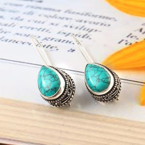 May include: A pair of silver drop earrings with turquoise teardrop-shaped stones. The stones are set in ornate, detailed silver settings. The earrings have a long, slender hook design.