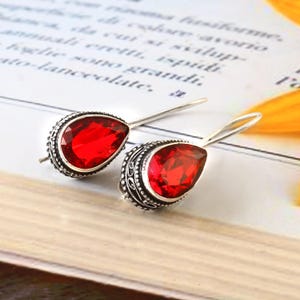 Peut inclure: Boucles d'oreilles pendantes en argent avec des pierres rouges en forme de goutte d'eau. Les pierres sont serties dans des montures argentées ornées. Les boucles d'oreilles sont posées sur une surface claire avec un texte flou en arrière-plan.