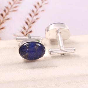 May include: Silver cufflinks featuring oval, deep blue lapis lazuli stones with gold inclusions. The stones are set in a silver bezel. The cufflinks are presented on a white surface.
