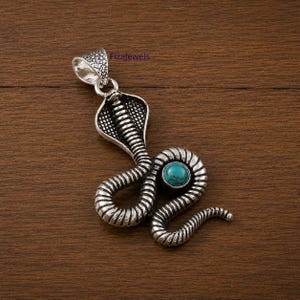 Puede incluir: Colgante plateado con forma de cobra con una piedra de color turquesa. El colgante tiene un diseño texturizado y el texto "Fizajewels" es visible.