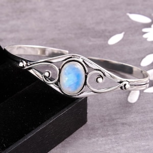 Könnte beinhalten: Ein silberfarbenes Armreif-Armband mit einem ovalen, hellblauen Edelstein. Das Armband hat ein offenes Design mit dekorativen Wirbeln und einer polierten Oberfläche. Der Edelstein ist der Mittelpunkt, eingebettet in eine detaillierte, verzierte Fassung.