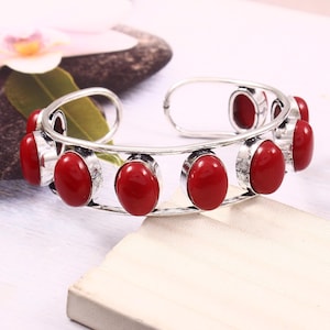Könnte beinhalten: Silberfarbenes Armreif-Armband mit ovalen, tiefroten Edelsteinen. Das Armband hat ein offenes Design, wobei die Edelsteine in einzelnen Fassungen oben angeordnet sind. Die Edelsteine sind gleichmäßig verteilt.