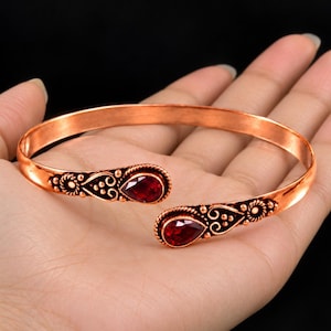 May include: A copper-toned open cuff bracelet featuring two teardrop-shaped red gemstones at the ends. The metal ends are decorated with intricate embossed scrollwork and small circular accents, creating a detailed metallic finish.