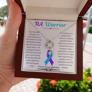 May include: A silver-toned necklace with a central knot pendant featuring a clear stone, displayed in a brown box. The card inside reads RA Warrior and includes a purple and blue awareness ribbon graphic with an inspirational message about resilience.