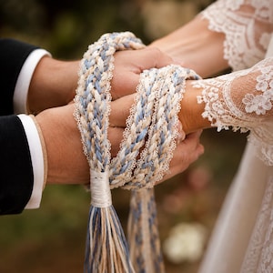 Könnte beinhalten: Ein Handfasting-Seil mit geflochtenem Design in Blau-, Beige- und Weißtönen. Das Seil ist an den Kanten mit zarter weißer Spitzenborte verziert und endet in langen, losen Quasten, die mit einem glatten weißen Faden zusammengebunden sind.