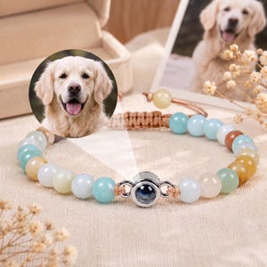 Könnte beinhalten: Ein Armband mit bunten Perlen in Blau, Grün und Gelb. Das Armband hat einen silberfarbenen Anhänger mit einem dunkelblauen Zentrum. Im Hintergrund ist ein Foto eines Golden Retrievers zu sehen.