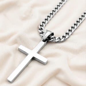 May include: A silver cross pendant necklace with a chunky curb chain. The cross is a simple, rectangular design. The necklace is displayed on a cream-coloured fabric.
