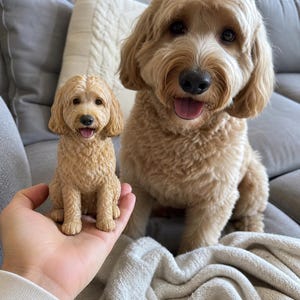 Può includere: Una piccola statuetta tenuta in mano di un cane color oro con pelo riccio, raffigurato in posizione seduta con la bocca leggermente aperta. La statuetta presenta texture dettagliate che imitano l'aspetto del pelo e i tratti del viso del cane.