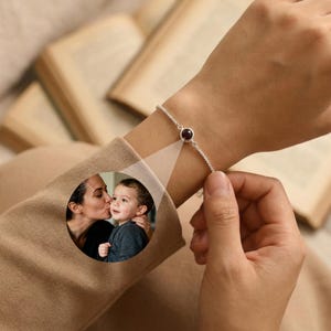 May include: A silver bracelet with a small, round, dark red gemstone. The bracelet is on a wrist, and a circular image of a mother kissing a child is projected onto the sleeve.