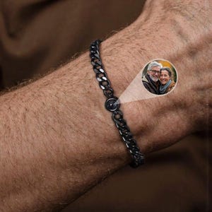 May include: A black chain bracelet with a circular charm, shown on a person's wrist. The charm appears to contain a small image of a couple. The bracelet is made of linked metal and has a dark, matte finish.