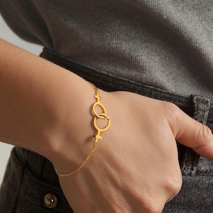 May include: A gold-colored bracelet featuring two interlocking Venus symbols, connected by a delicate chain. The bracelet is worn on a wrist, with the hand resting on a gray surface. The Venus symbols are a prominent design element.