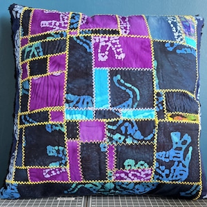 May include: Square cushion with a patchwork design. The cushion features a mix of purple, blue, and black fabrics, stitched together with yellow thread. The fabric has a batik-like pattern.