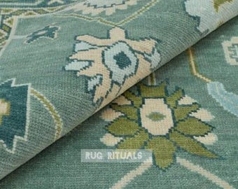 May include: A teal area rug featuring a repeating geometric floral pattern in shades of sage green, cream, and light blue. The rug is folded over to reveal the underside, and the text RUG RITUALS is printed in white on the surface of the fabric.