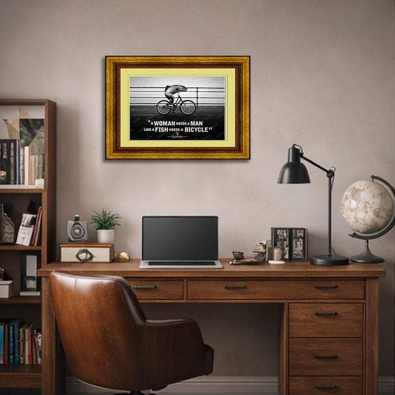 May include: Framed artwork with a gold and brown border. The image features a black and white illustration of a fish riding a bicycle. The text reads: "A woman needs a man like a fish needs a bicycle."