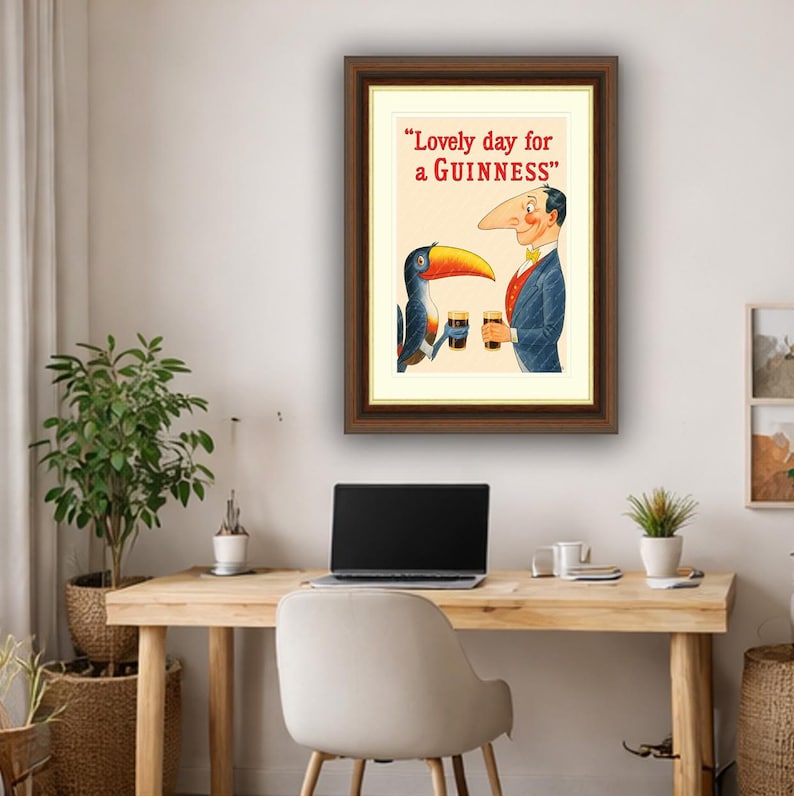 Puede incluir: Anuncio vintage enmarcado de Guinness con un tuc&aacute;n de dibujos animados y un hombre con una nariz larga, ambos sosteniendo pintas de cerveza negra. El texto dice "Lovely day for a Guinness" en letras rojas. La obra de arte est&aacute; en un marco de madera marr&oacute;n.