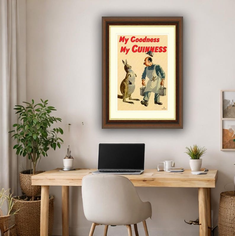 K&ouml;nnte beinhalten: Gerahmte Vintage-Guinness-Werbung mit einem K&auml;nguru und einem Mann mit Eimern. Der Text "My Goodness My Guinness" ist rot dargestellt. Das Kunstwerk befindet sich in einem braunen Rahmen und wird an einer hellen Wand &uuml;ber einem Holzschreibtisch gezeigt.