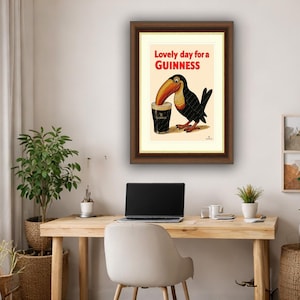 May include: Framed vintage Guinness advertisement featuring a toucan drinking a pint of Guinness. The text "Lovely day for a Guinness" is in red. The artwork is in a brown frame, displayed on a light-colored wall above a wooden desk.