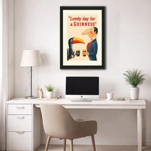 Puede incluir: Anuncio vintage de Guinness enmarcado con un tuc&aacute;n de dibujos animados y un hombre con una nariz grande, ambos sosteniendo vasos de cerveza negra. El texto dice "Lovely day for a Guinness" en rojo y negro. La obra de arte est&aacute; en un marco negro.