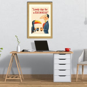 Puede incluir: Un anuncio vintage enmarcado de Guinness stout, con un tuc&aacute;n de dibujos animados y un hombre. El texto dice: "Lovely day for a Guinness". La imagen se muestra encima de un escritorio de madera con un port&aacute;til y una taza roja.