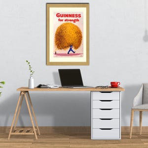 May include: Framed vintage Guinness advertisement. The print features a man in blue carrying a large, golden-yellow sphere with the text "Guinness for strength" in red. The artwork is displayed above a desk with a laptop and a red mug.