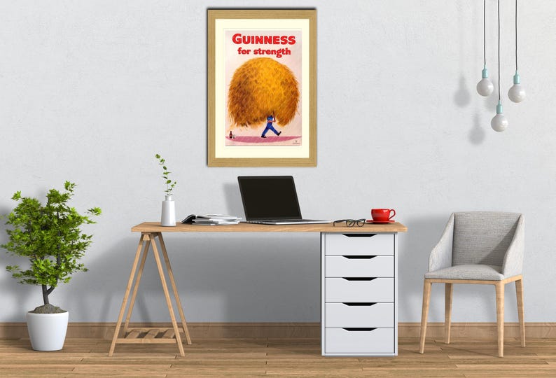 May include: Framed vintage Guinness advertisement. The print features a man in blue carrying a large, golden-yellow sphere with the text "Guinness for strength" in red. The artwork is displayed above a desk with a laptop and a red mug.