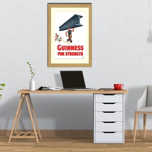 May include: Framed vintage Guinness advertisement. The print features a man carrying a large steel beam, with the text "Guinness For Strength" in red. The image is displayed on a light-colored wall above a wooden desk.
