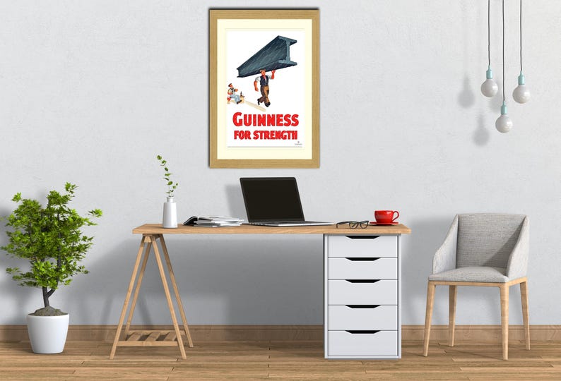 May include: Framed vintage Guinness advertisement. The print features a man carrying a large steel beam, with the text "Guinness For Strength" in red. The image is displayed on a light-colored wall above a wooden desk.