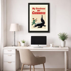 May include: Framed vintage Guinness advertisement featuring a black seal balancing a pint of Guinness. The poster has the text "My Goodness My Guinness" in red and black. A man in a green uniform is running towards the seal.