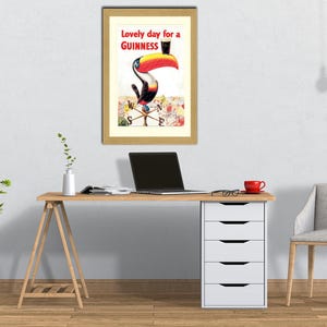 Puede incluir: Un anuncio vintage enmarcado de Guinness con un tuc&aacute;n con sombrero de copa. El texto dice "Lovely day for a Guinness". La obra de arte est&aacute; colgada sobre un escritorio de madera con un port&aacute;til y una taza roja.