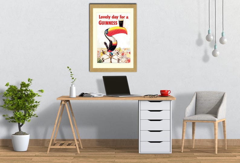 Puede incluir: Un anuncio vintage enmarcado de Guinness con un tuc&aacute;n con sombrero de copa. El texto dice "Lovely day for a Guinness". La obra de arte est&aacute; colgada sobre un escritorio de madera con un port&aacute;til y una taza roja.