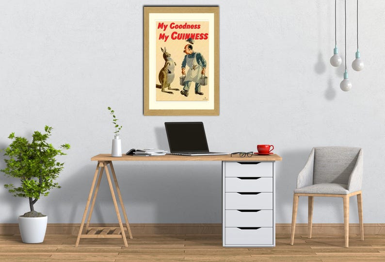 K&ouml;nnte beinhalten: Ein gerahmtes Vintage-Guinness-Poster mit einem K&auml;nguru und einem Mann in einer Arbeitssch&uuml;rze. Der Text "My Goodness My Guinness" ist in Rot und Schwarz dargestellt. Das Poster befindet sich auf einem Schreibtisch mit einem Laptop, einer roten Tasse und einem Stuhl.