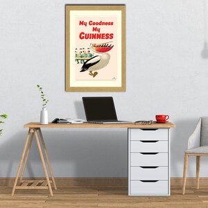 May include: A framed vintage advertisement for Guinness stout. The artwork features a pelican carrying a tray of drinks, with the text "My Goodness My Guinness" in red and white. The frame is a light wood color.