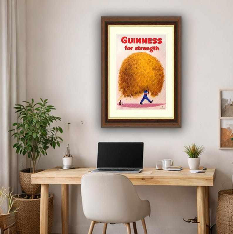 May include: Framed vintage Guinness advertisement. The artwork features a man in blue carrying a large, golden-brown bundle. The text "GUINNESS for strength" is in red at the top. A bottle is on the ground near the man.
