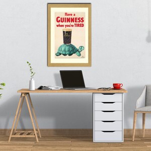 K&ouml;nnte beinhalten: Gerahmte Vintage-Guinness-Werbung. Das Kunstwerk zeigt ein Pint Guinness auf einer Schildkr&ouml;te mit dem Text "Have a Guinness when you're TIRED". Der gerahmte Druck befindet sich &uuml;ber einem Holztisch mit einem Laptop und einer roten Kaffeetasse.