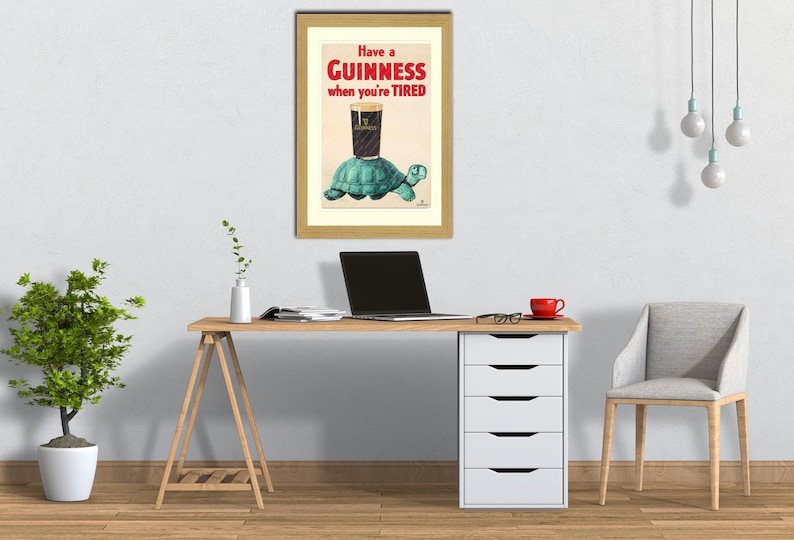 K&ouml;nnte beinhalten: Gerahmte Vintage-Guinness-Werbung. Das Kunstwerk zeigt ein Pint Guinness auf einer Schildkr&ouml;te mit dem Text "Have a Guinness when you're TIRED". Der gerahmte Druck befindet sich &uuml;ber einem Holztisch mit einem Laptop und einer roten Kaffeetasse.