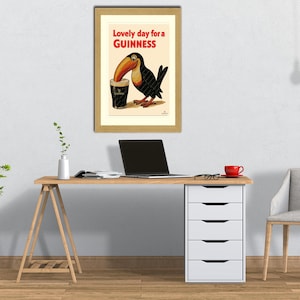 May include: Framed vintage Guinness advertisement featuring a toucan drinking a pint of Guinness. The poster has the text "Lovely day for a Guinness" in red and black. The framed poster is on a white wall above a wooden desk.