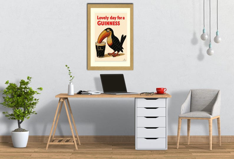 May include: Framed vintage Guinness advertisement featuring a toucan drinking a pint of Guinness. The poster has the text "Lovely day for a Guinness" in red and black. The framed poster is on a white wall above a wooden desk.