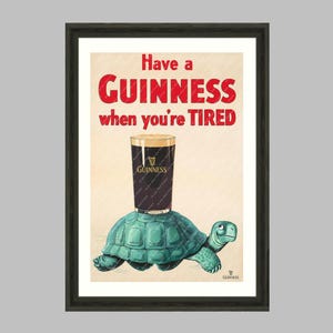 K&ouml;nnte beinhalten: Gerahmter Wandkunstdruck mit einem Guinness-Pint, der auf einer t&uuml;rkisfarbenen Schildkr&ouml;te balanciert. Der Text lautet "Have a Guinness when you're TIRED" in Rot und Wei&szlig;. Das Kunstwerk stammt aus "The Wall Art Parlour Collection".