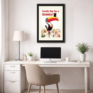 Puede incluir: Anuncio vintage enmarcado de Guinness con un tuc&aacute;n sosteniendo una pinta de Guinness. El tuc&aacute;n tiene un pico grande rojo y amarillo. El texto en la parte superior dice "Lovely day for a Guinness". La obra de arte est&aacute; sobre un fondo crema.